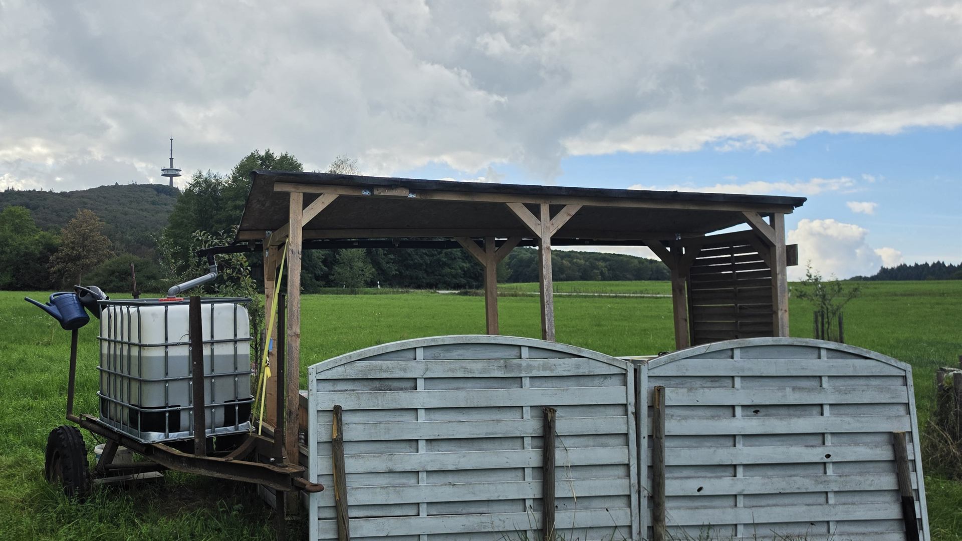 Bienenstand am Fuße des Dünsbergs: ein hölzerner Unterstand mit Bienenbeuten auf einer grünen Wiese, davor ein Wassertank auf einem Anhänger, im Hintergrund der bewaldete Dünsberg mit seinem Sendeturm unter einem wolkigen Himmel